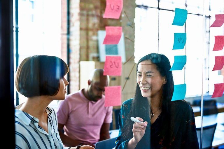 Two people discussing in front of a glass wall with sticky notes, one smiling. A person sitting on a table in the background.