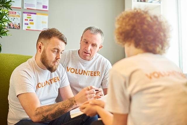 Three people wearing Volunteer shirts are seated indoors, engaged in conversation about charity work. A man with a beard is writing in a notebook. The background shows colorful charts on the wall and a green couch, illustrating their plans for an upcoming event.