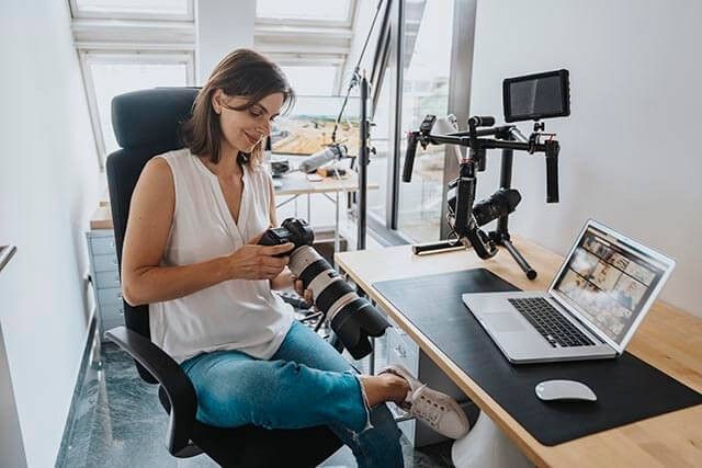 A person sits at a desk in a home office, examining a camera with a large lens. A laptop and camera equipment, including a stabiliser, are on the desk. The setting is bright with natural light from windows.