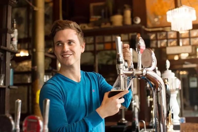 A bartender is smiling while pouring a drink from a tap at a bar with shelves and bottles the background.
