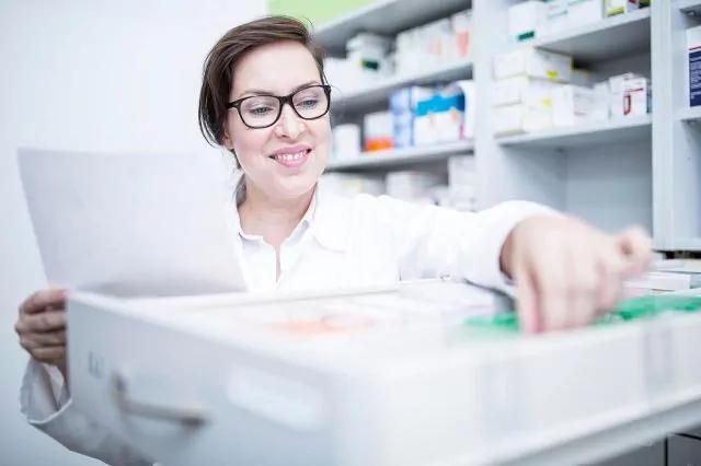 A woman wearing glasses and a white coat stands in front of shelves with pharmaceutical products, holding a sheet of paper in her left hand. As she reaches into an open drawer with various items using her right hand.