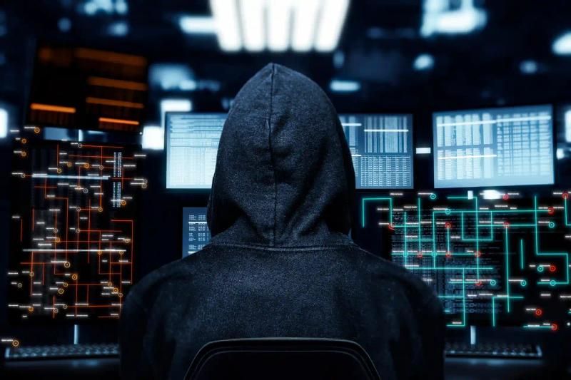 A person in a hoody facing monitors displaying digital data, conducting a cyber attack in a dark room