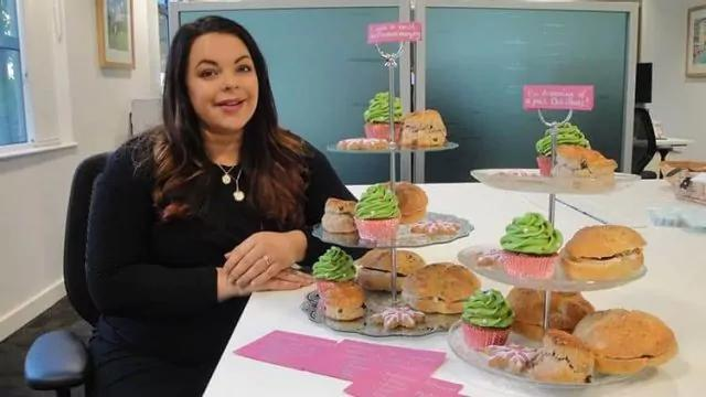 A woman seated at a table with multiple trays of baked goods, including scones and green frosted cupcakes. She is smiling, and there are small pink signs and cards on the table next to her. The setting appears to be an office or meeting room.