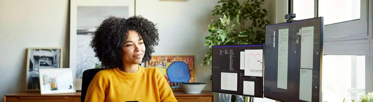 A woman seated at a desk, focused on her computer screen, surrounded by office supplies and a window in the background.