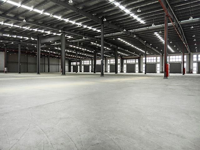 Spacious, empty warehouse with concrete floor, metal beams, and natural light coming in from big windows.