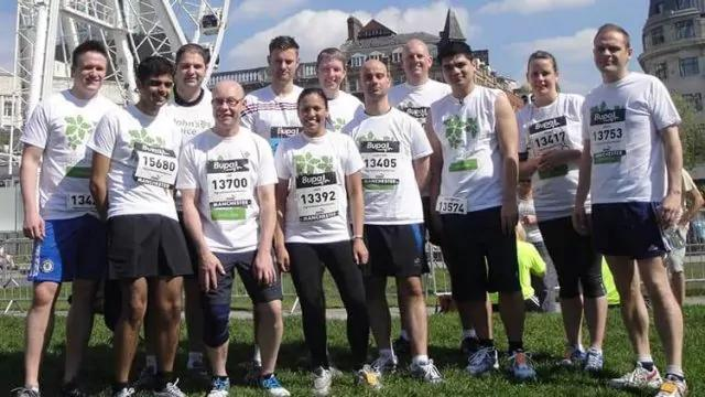 A group of twelve people in athletic gear stand together on grass in front of a Ferris wheel. They are wearing numbered race bibs, indicating participation in a running event. The background shows buildings and a clear sky.