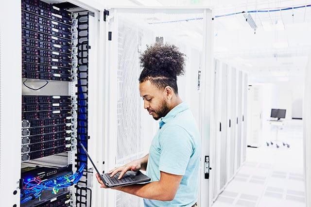 A person with curly hair in a bun is standing in a server room, using a laptop. They are in front of a server rack filled with various networking equipment and cables. The room is well-lit, with multiple server cabinets visible.