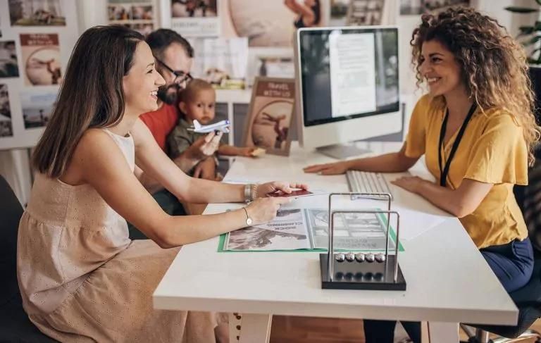 Couple with a baby consulting a travel agent in an office, with travel posters in the background.