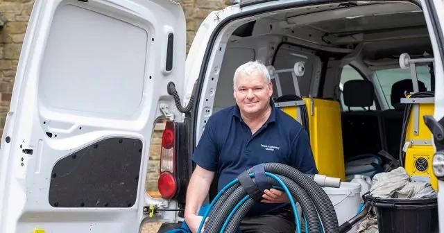 Man sitting at the back of an open white van with cleaning supplies, holding a large hose.
