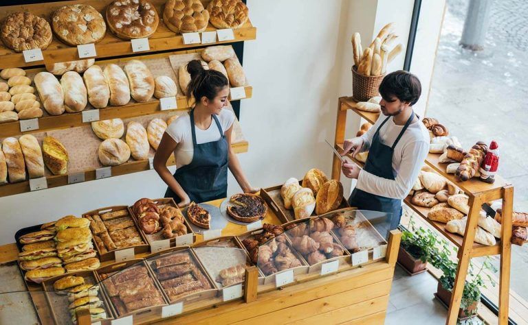 Two bakery workers behind a counter with breads and pastries, one using a tablet while the other arranges items.