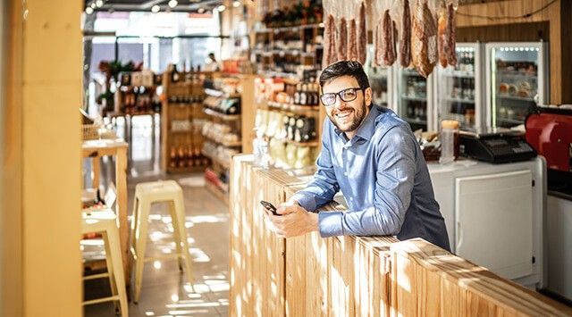 Man leaning on a counter in a delicatessen shop, smiling and holding a smartphone.