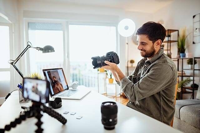 A man sits at a desk in a well-lit room, holding a camera. A laptop and tripod-mounted smartphone are on the desk. The laptop displays a video call with another person. Various camera lenses and accessories are also visible.