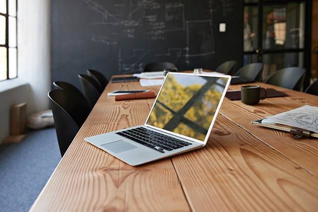 An open laptop on top of a wooden office desk, with trees and windows reflected on the turned-off screen.