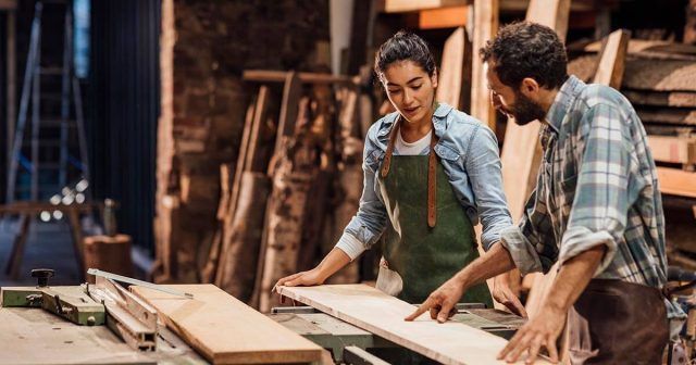 Two people in a woodworking shop discussing a wooden plank, surrounded by tools and wood.