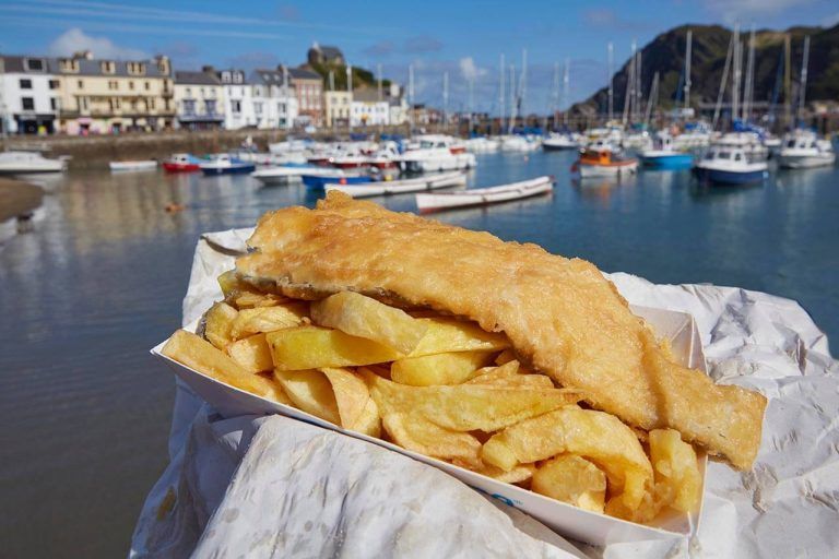 Fish and chips served on paper near a harbor, with boats moored in the background.