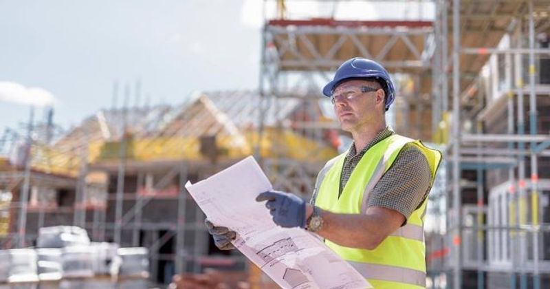 A construction worker, clad in a yellow safety vest, blue hard hat, and gloves, examines blueprints at a bustling site. Scaffolding rises against the clear sky.