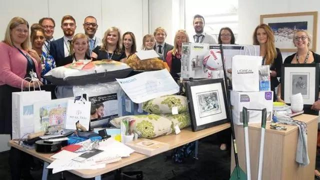 A group of people is standing behind a table filled with various items, including framed pictures, pillows, bags, and gift boxes. The setting appears to be an office with a window and some artwork on the walls.