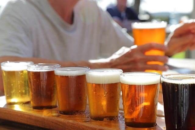 A wooden tray holds six glasses of beer in varying shades from light to dark. In the background, a person wearing a light-colored shirt holds a pint of amber beer. The setting appears to be an outdoor seating area on a sunny day.