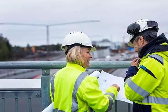 Two construction workers discussing plans on a bridge, with a construction site and road in the background.