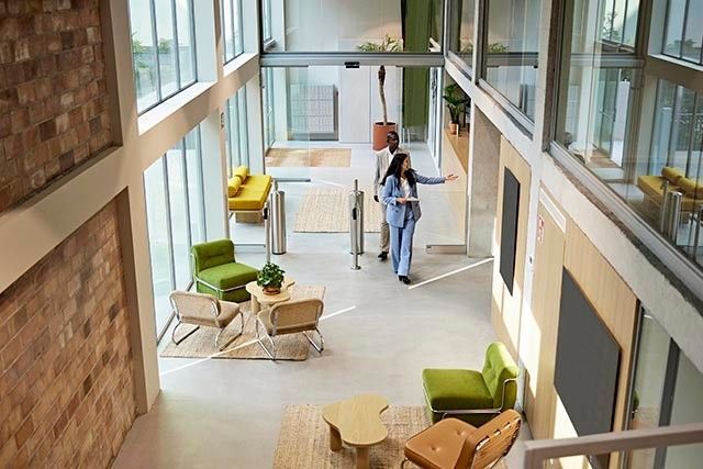 Modern communal office lobby with natural light, and chairs. A person appears to be giving a tour to another one.