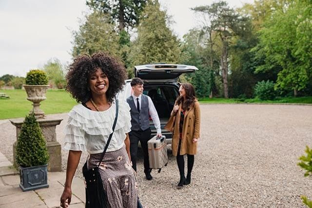 Three people standing in a hotel car park. A woman is smiling while an employee is helping with the suitcase.