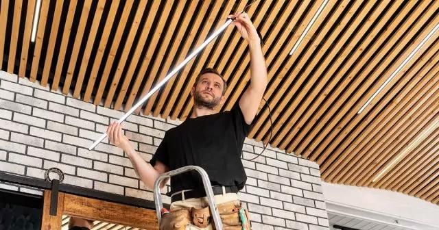 Man installing a fluorescent light bulb on a ladder in a room with a brick wall and wooden ceiling, wearing a tool belt.