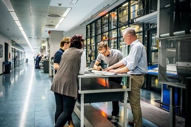 Four people stand around a table in a brightly lit hallway, engaged in discussion. They are examining engineering blueprints. Shelves filled with books or files line the wall in the background.