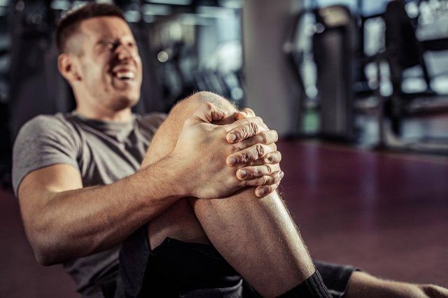 Man lying on the gym floor, holding his injured knee, in pain, with exercise equipment around.