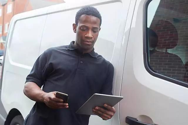 Man in black polo standing beside a white van, holding a tablet and phone near a brick building.