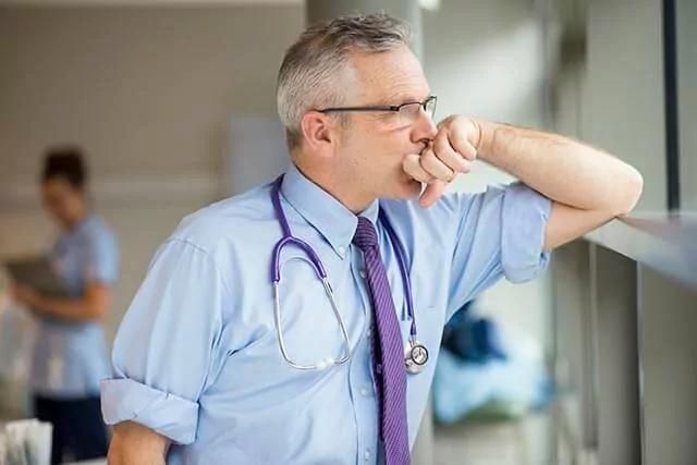 A male doctor in a blue shirt, and tie, with a stethoscope leans against a window, looking thoughtfully outside.