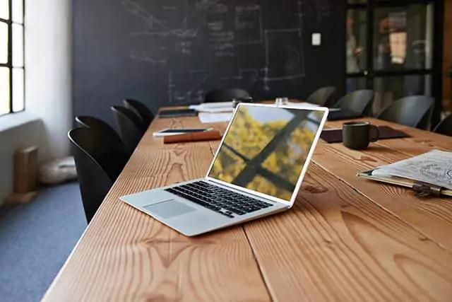 An open laptop on top of a wooden office desk, with trees and windows reflected on the turned-off screen.