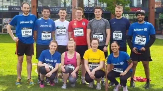 A group of eleven people, comprising both men and women, are standing together on grass. They are wearing athletic clothing and race numbers. A building with shop signs is visible in the background.