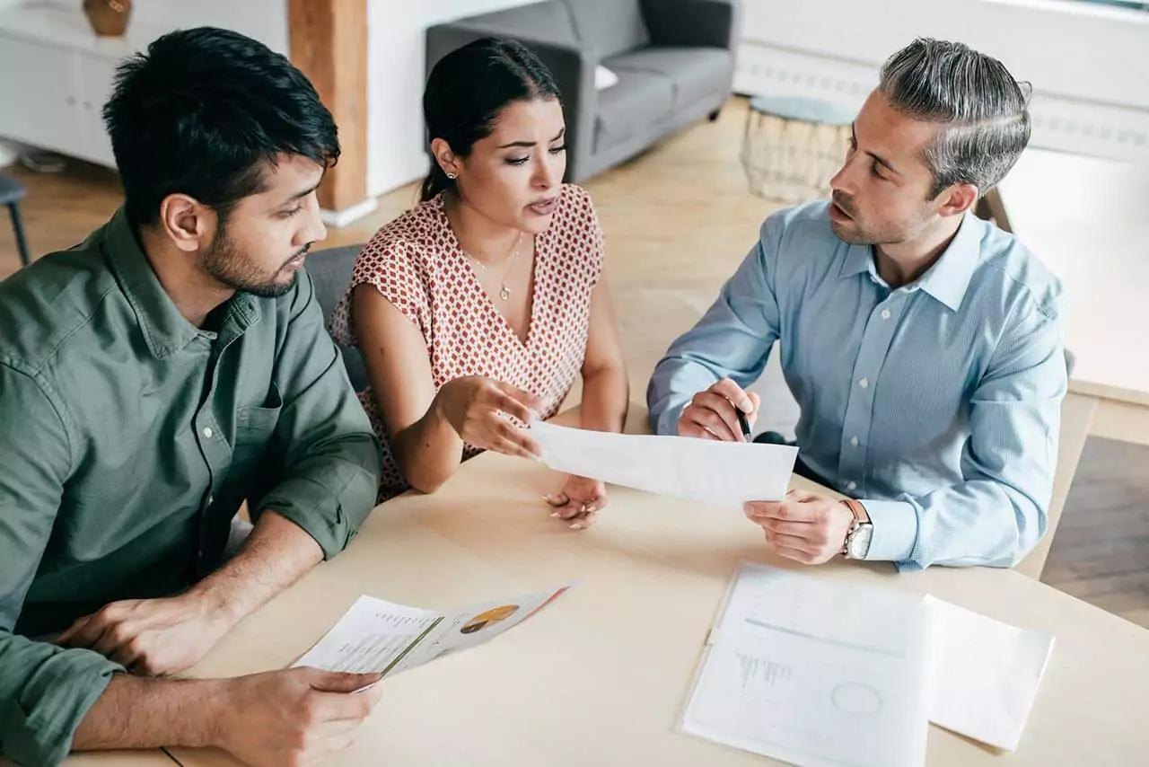 Group of professionals in a meeting room discussing documents, sitting around a wooden table.