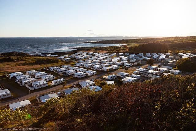 A scenic view of a coastal caravan park with numerous white caravans arranged in neat rows. The ocean is visible in the background under a clear sky, while the foreground features greenery and shrubs. The sunlight casts a warm glow over the area.