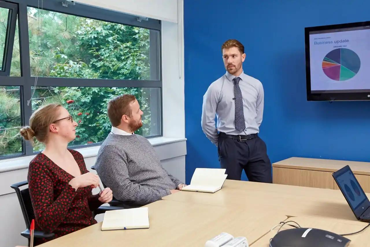 Employee in a business meeting, presenting a business update to a seated audience with a laptop on the table.