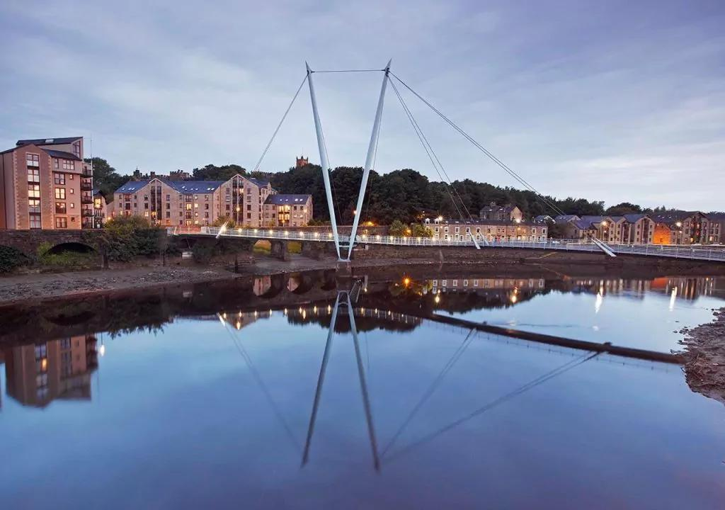 Modern footbridge over the River Lune in Lancaster, with residential buildings and a cloudy evening sky.