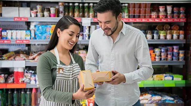Woman and man smiling in a grocery store aisle, holding packaged food items with stocked shelves.