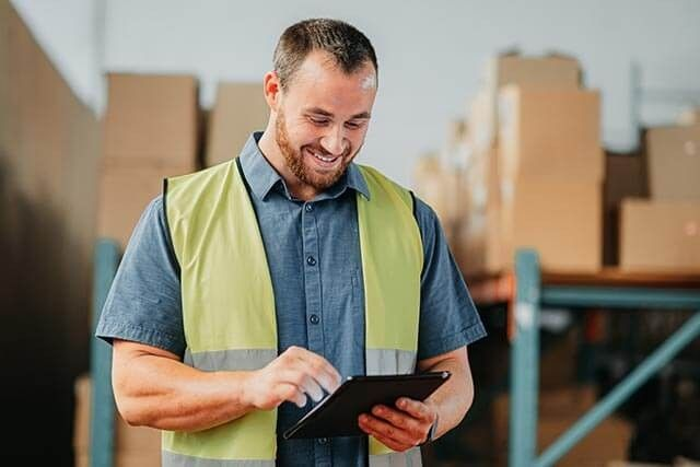 Man in a warehouse using a tablet, with stacks of boxes and metal shelving in the background.