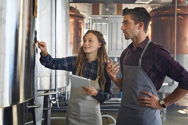Two people in aprons stand in a brewery. One holds a tablet and points at a metal container, while the other stands with a hand on their hip. Industrial equipment is visible in the background.