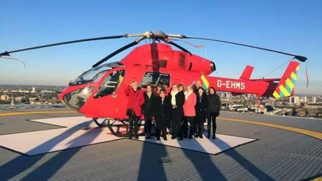 A group of people stand in front of a red helicopter on a rooftop helipad. The helicopter has the registration G-EHMS. The background shows a cityscape under a clear blue sky.