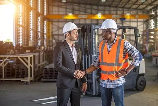 Two men in a warehouse shaking hands, one in a suit, the other in safety gear, with a forklift and stacked materials behind.