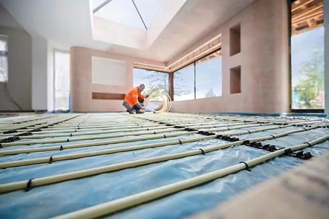 Worker installing underfloor heating in a spacious room with a skylight. The floor is covered with blue sheeting and pipes.