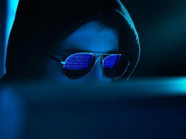 Hacker in a dark hoodie working on a laptop in a dimly lit room with code reflected on his black sunglasses.