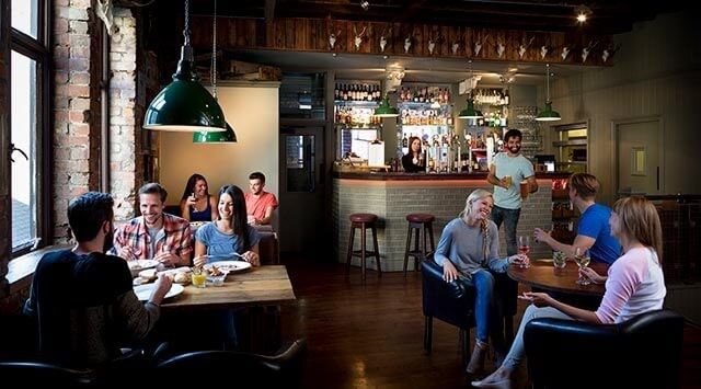 Cozy café scene with people enjoying food and drinks, wooden décor, and a bartender serving behind the bar.