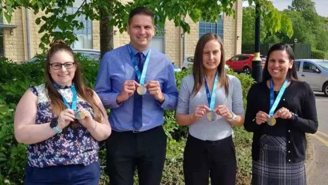 Four people stand outdoors holding medals. They are under a tree, dressed in business-casual attire, and smiling for the photo. A building, greenery, and parked cars are in the background.