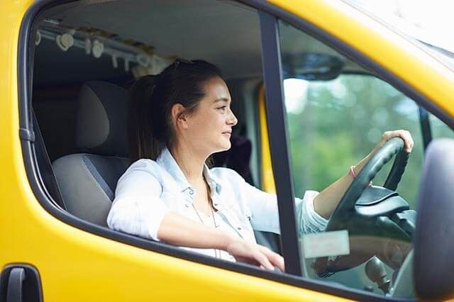 A person with long hair is sitting in the drivers seat of a yellow vehicle, looking forward. One hand is on the steering wheel, and the other rests near the window. The background is blurred greenery.