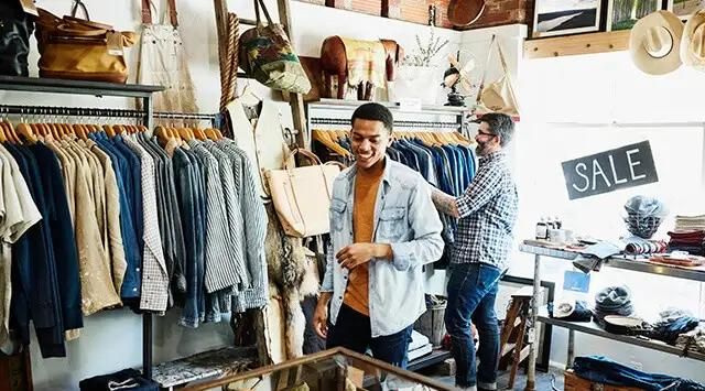 Customers are browsing clothes in a store with racks of shirts and jackets. A sign on the window reads 'SALE'.