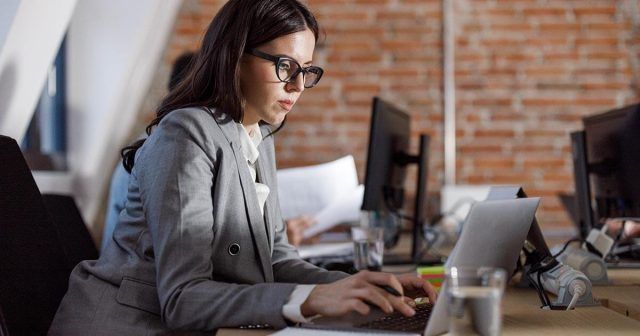 A woman in a grey business suit focused on her laptop, engaged in work at a modern office setting.