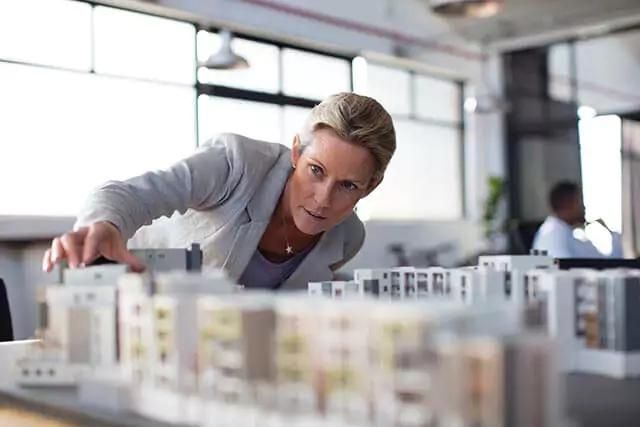 A person in a gray blazer examining a scale model of a building in an office with large windows.