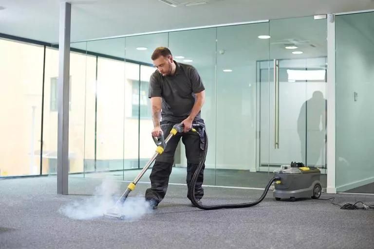 Man cleaning a grey carpet with a steam cleaner in a modern office, steam rising.
